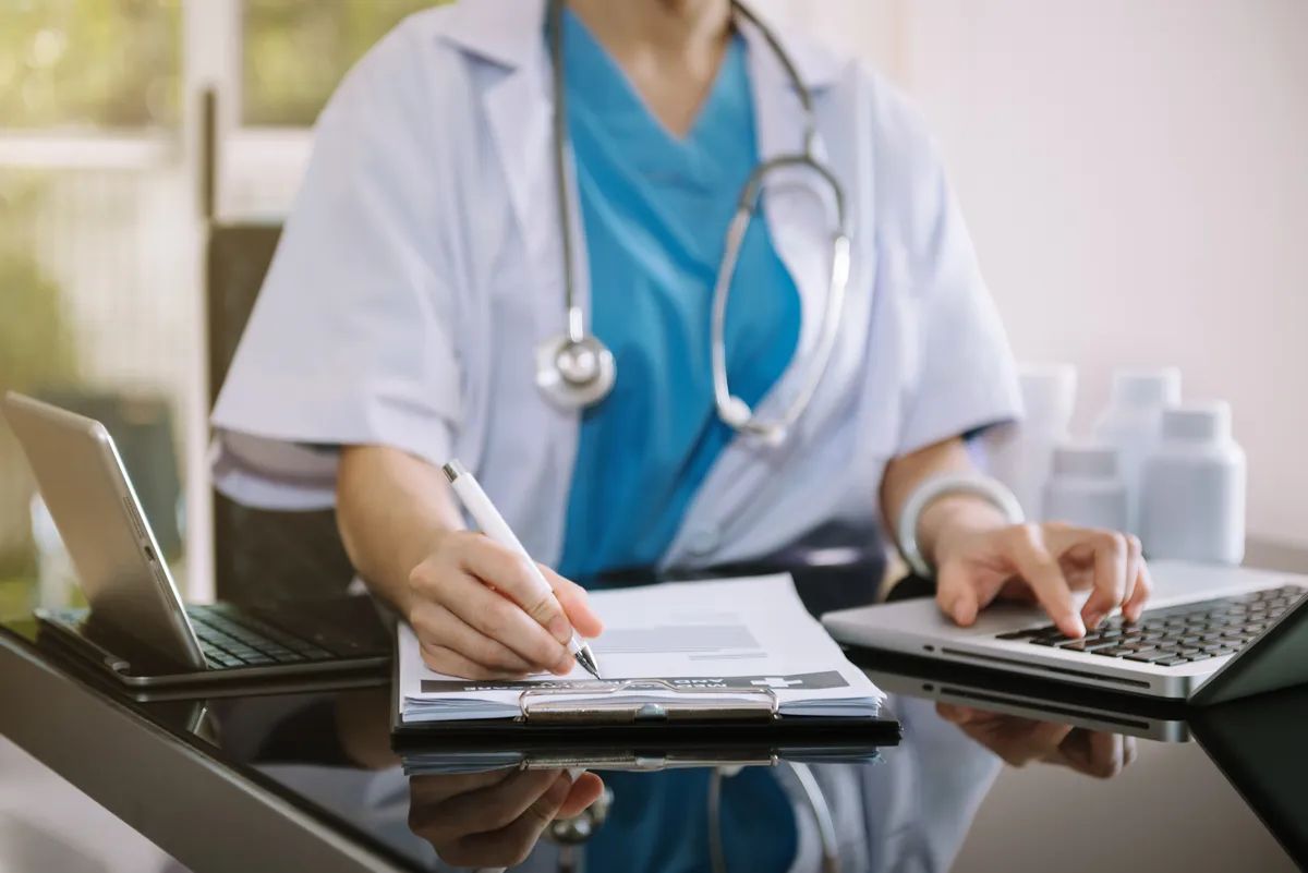 Clinician in scrubs reviewing patient records beside a laptop in a clinical setting