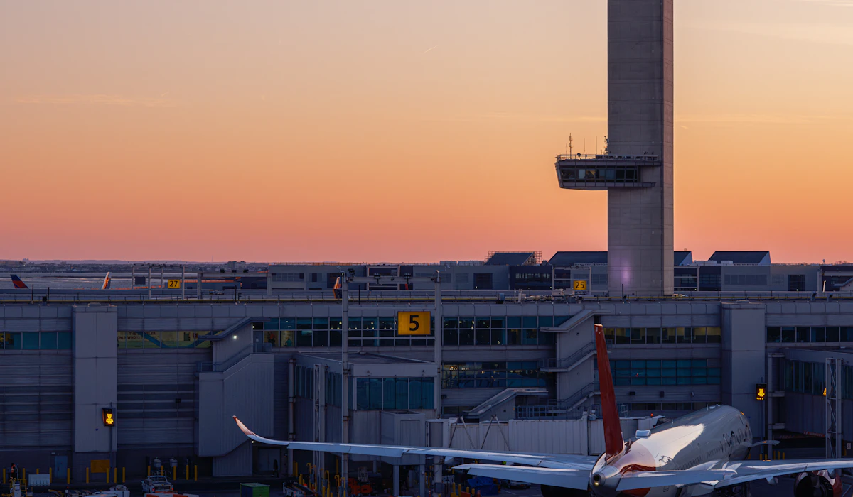 An airport control tower rising above a terminal building at sunset, with an airliner parked at a gate below and jet bridges extending along the pier