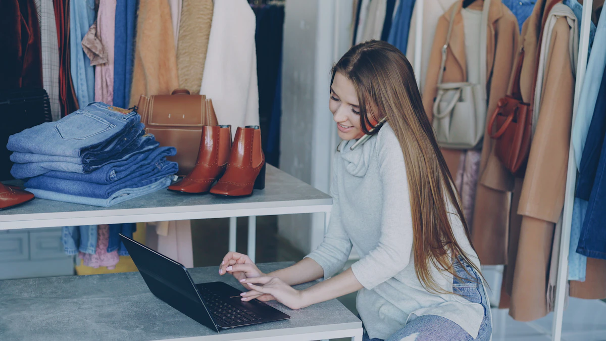 A boutique owner sitting at a grey counter working on a laptop with a phone to her ear, surrounded by rails of clothing, folded jeans and handbags