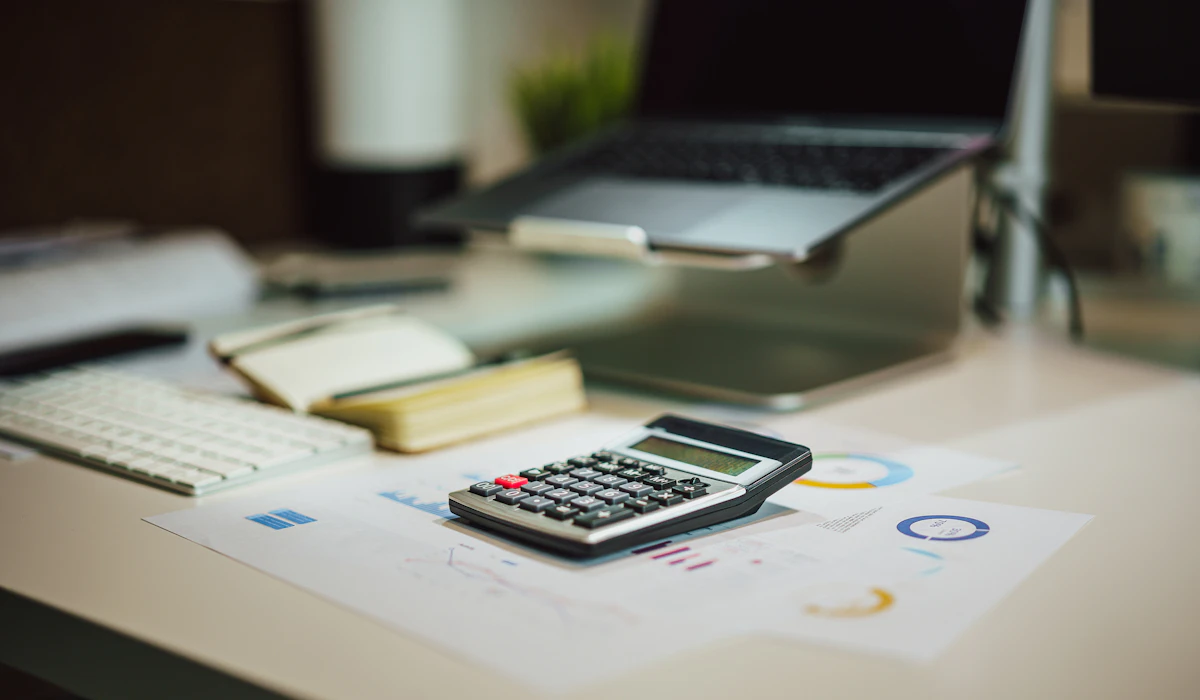 A calculator resting on printed financial charts next to a laptop