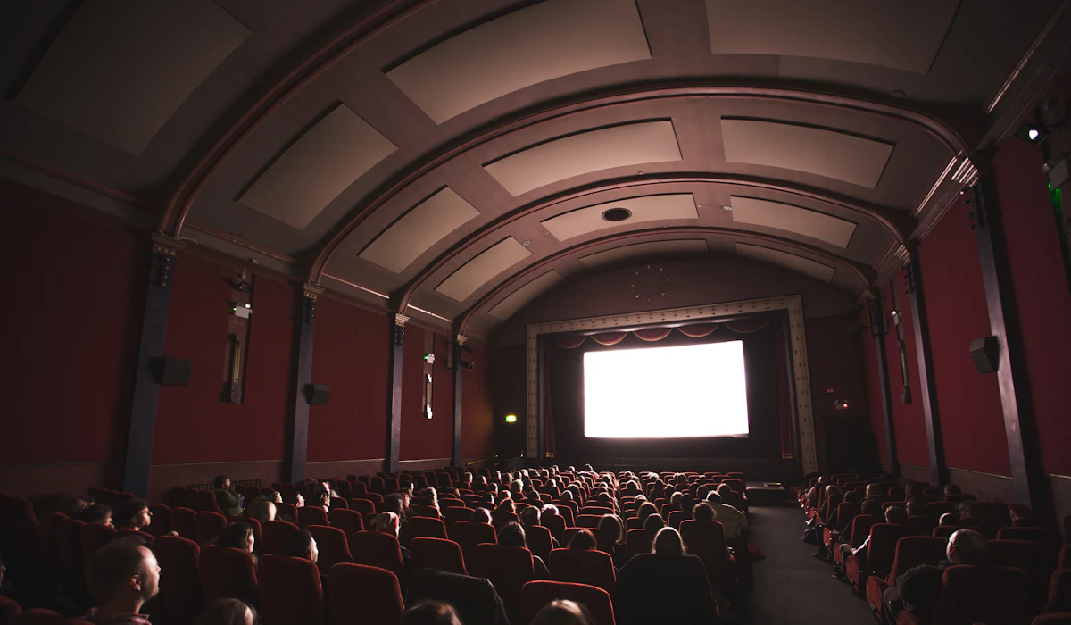 An audience in a dark cinema with red velvet seats facing a bright screen
