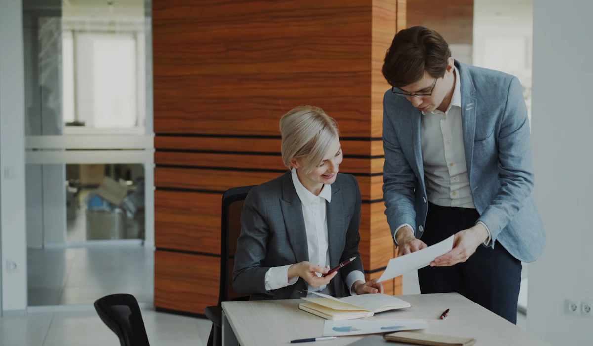 Two colleagues in an office, one seated and one standing, reviewing a printed document together beside an open notebook