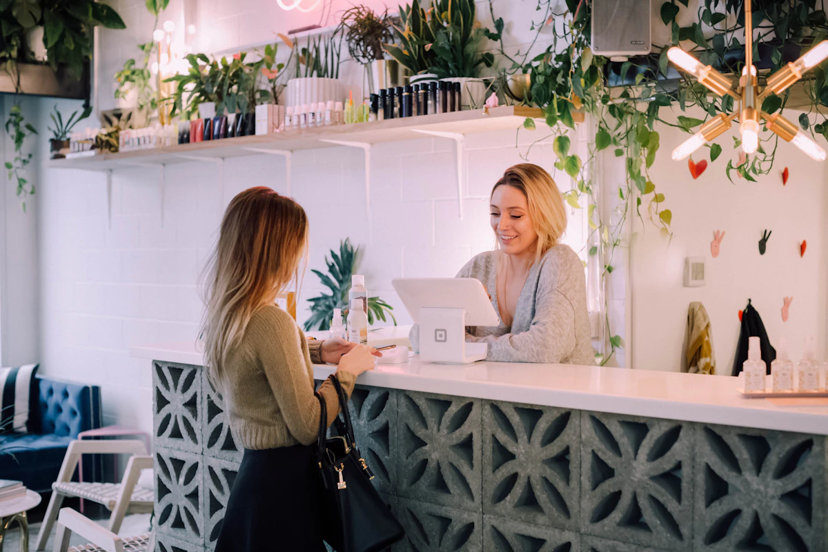 A customer standing at a plant-filled shop counter paying a smiling assistant at a tablet till