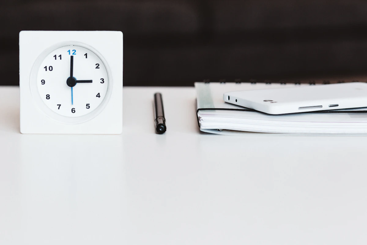 A white desk clock, a pen and a notebook on a plain white desk