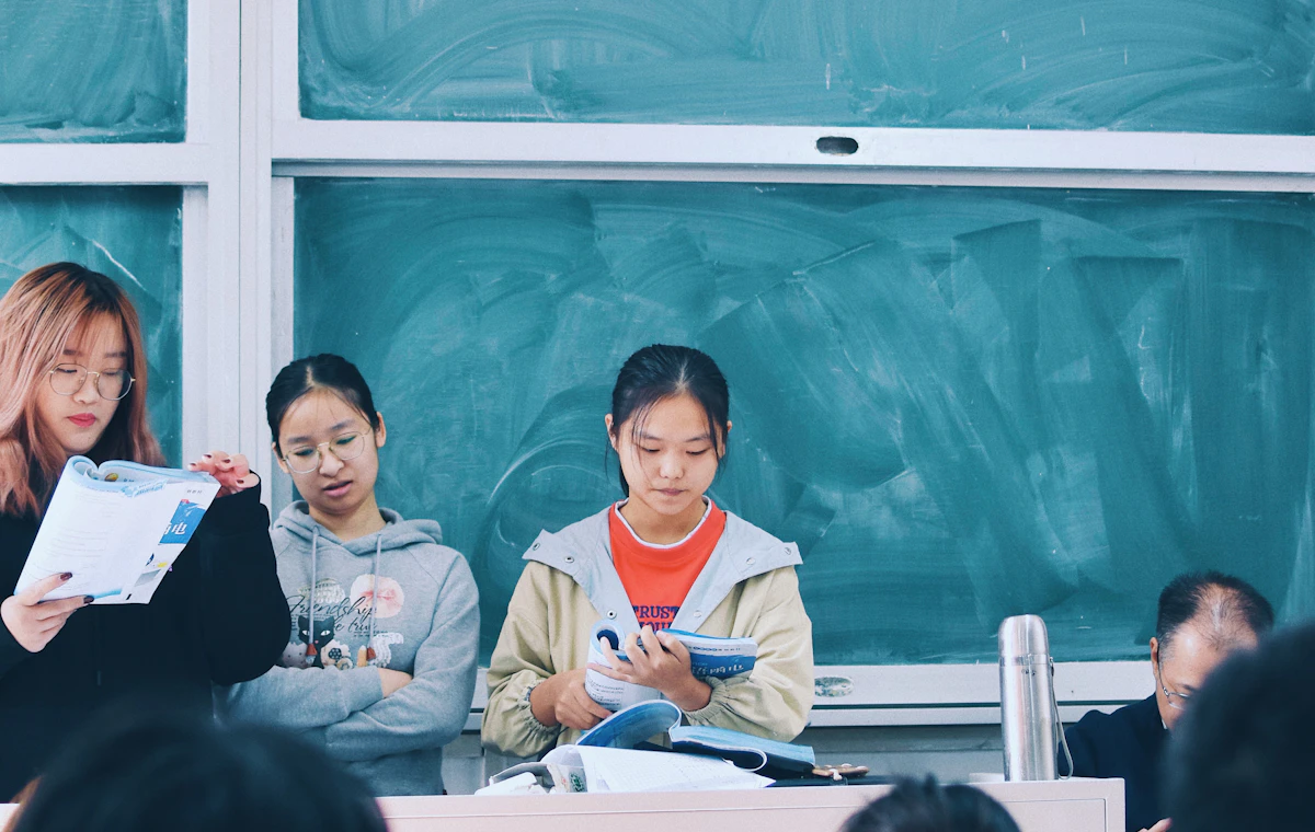 Three students standing at the front of a classroom reading from English workbooks, with a chalkboard behind them and classmates watching from their desks