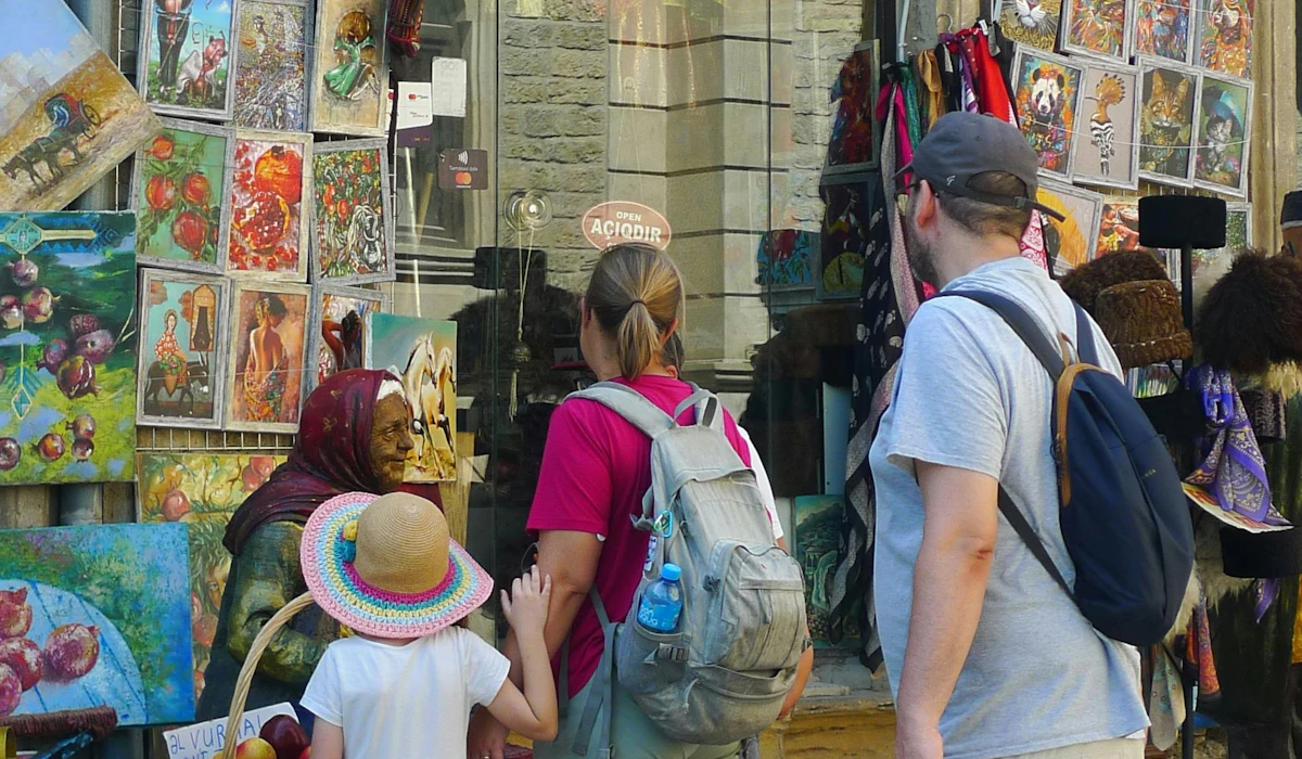 A family with backpacks and a child in a sun hat browsing paintings and scarves outside a souvenir shop while the vendor sits among the canvases