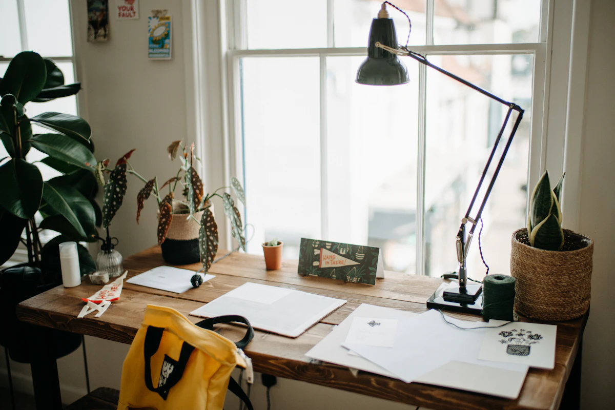 A freelancer's wooden desk by a window with papers, a lamp and plants
