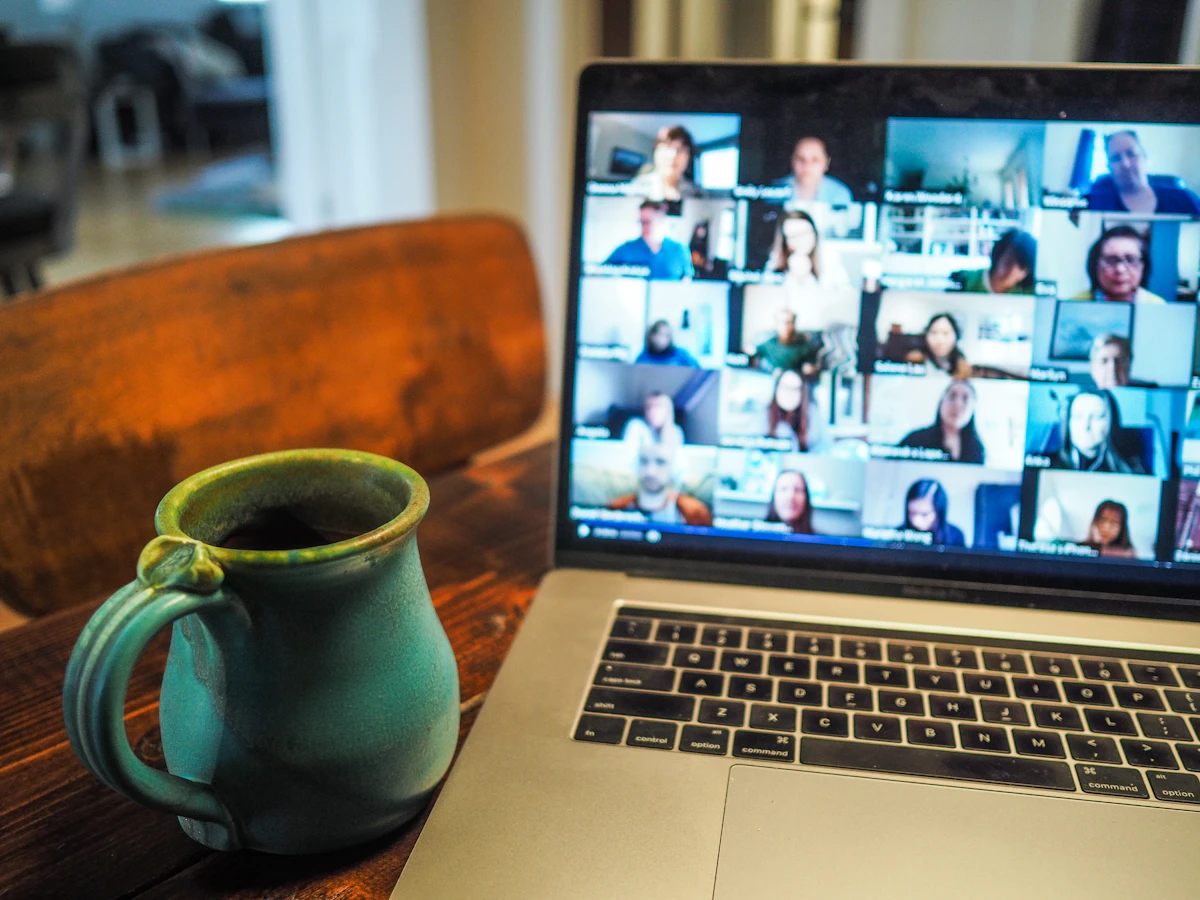 A laptop on a home dining table showing a video call with dozens of participants in a grid, next to a green ceramic coffee mug
