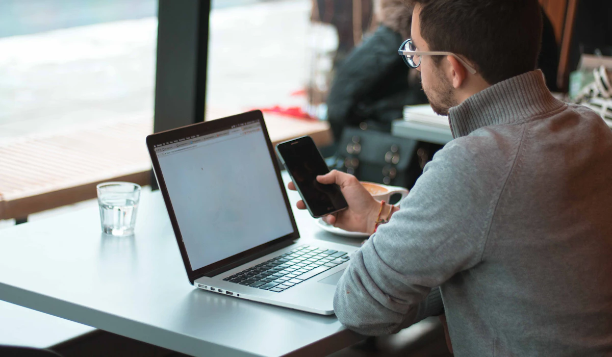 A man checking his phone next to an open laptop by a window