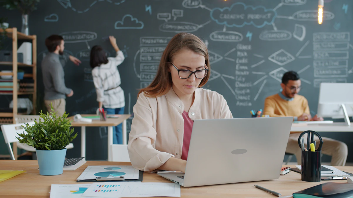 A marketer working at a laptop with printed charts on the desk, colleagues mapping ideas on a chalkboard wall behind her