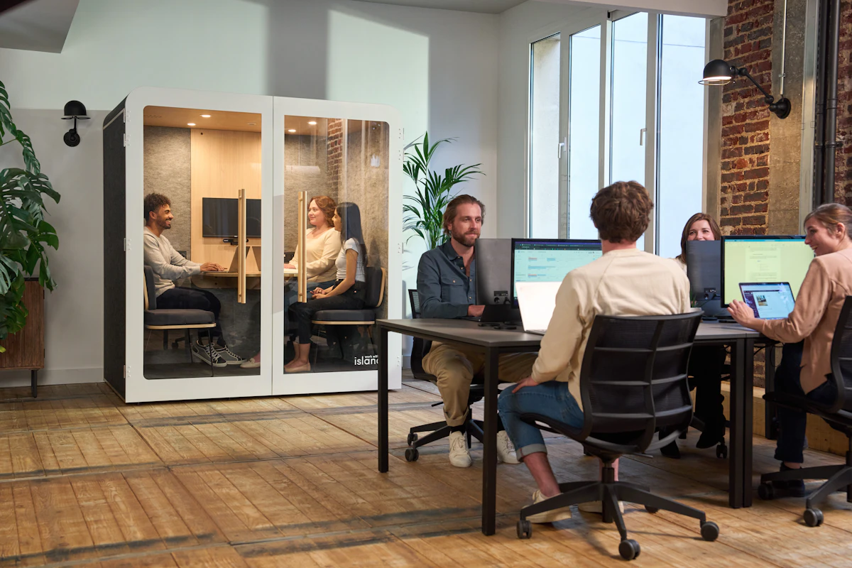An open-plan office with a glass soundproof meeting pod on one side and colleagues talking at desks with monitors on the other