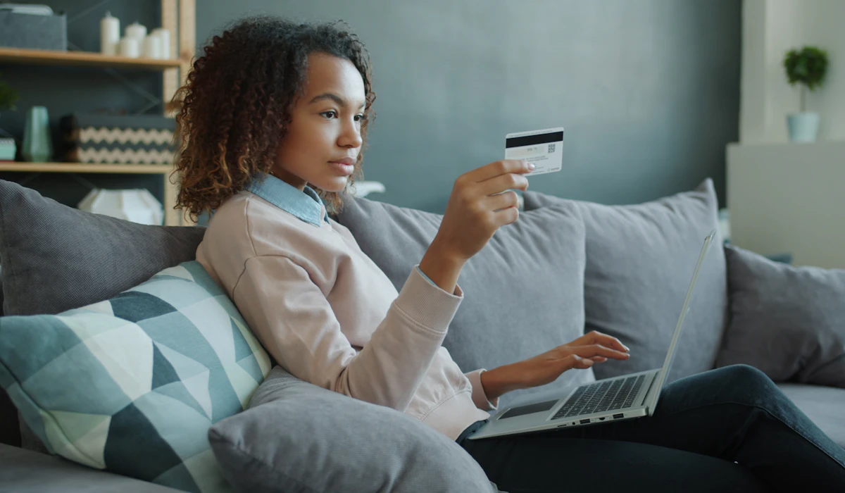 A woman sitting on a sofa with a laptop on her knees, holding a payment card up and reading the screen before deciding whether to use it