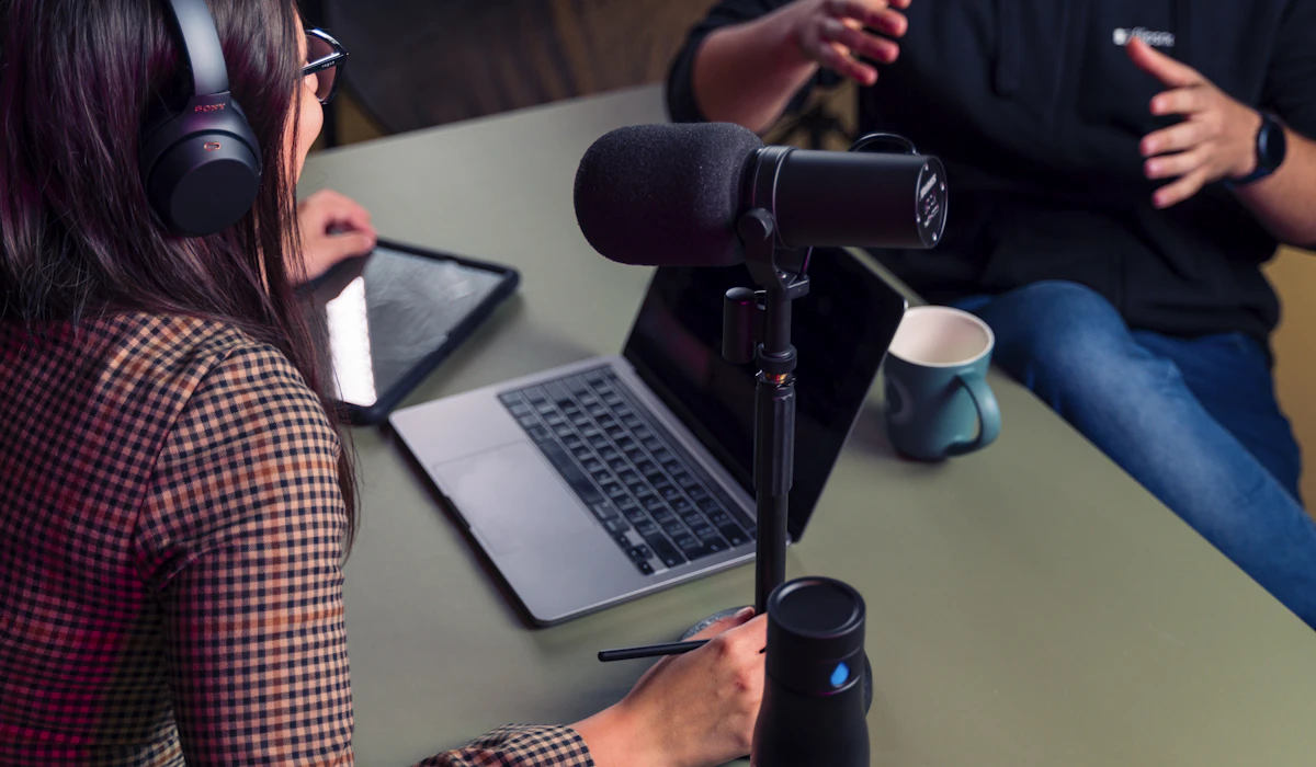 A podcast host in over-ear headphones taking notes at a laptop behind a studio microphone, with a guest gesturing across the table