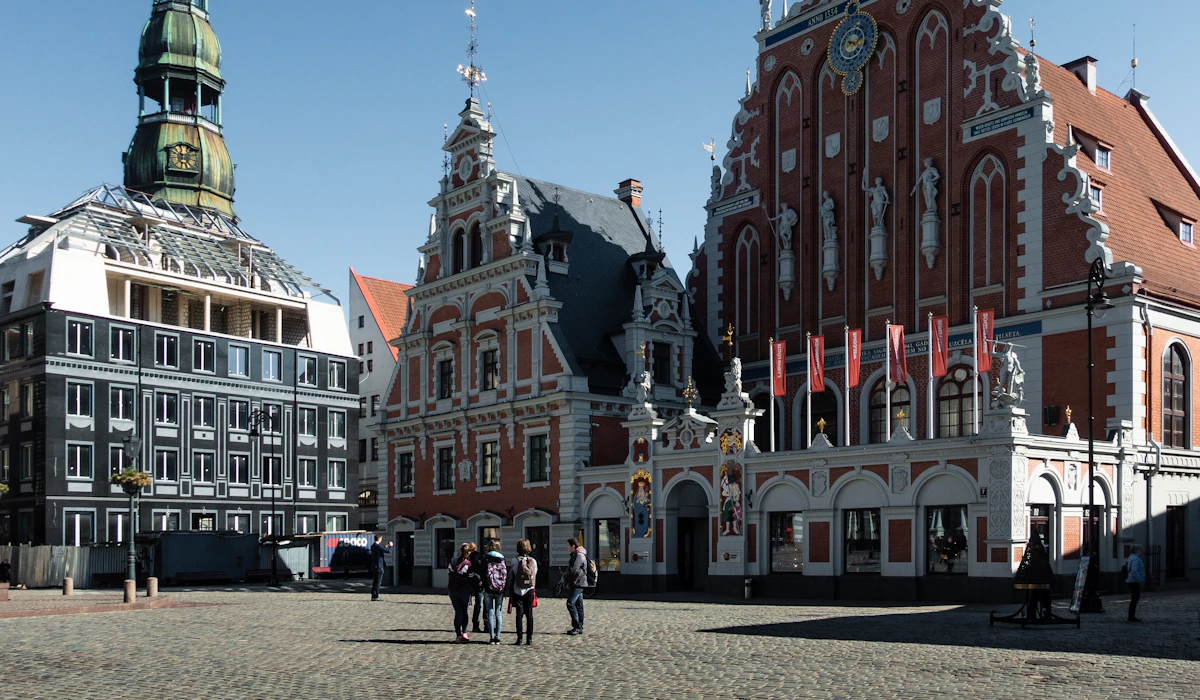 An ornately decorated red-brick building with a stepped gable and carved stone figures on a cobbled square in Riga, a green copper church spire rising behind it
