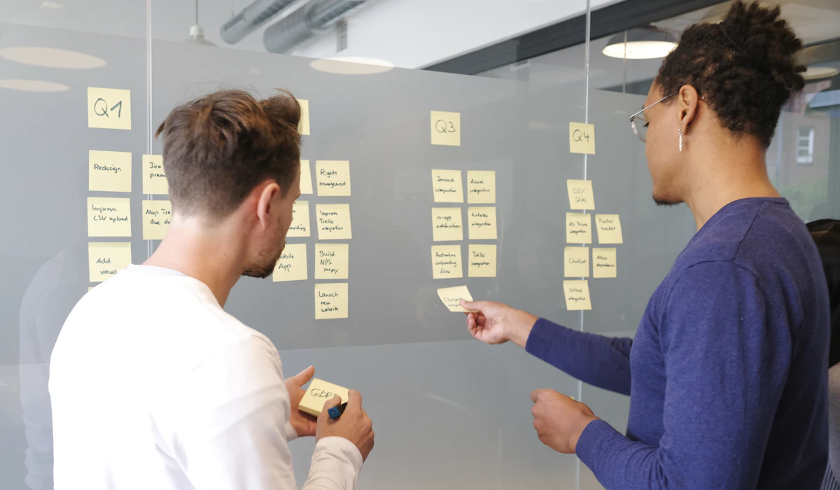 Two colleagues standing at a glass wall covered in yellow sticky notes grouped under handwritten quarter headings, one holding a note and a marker