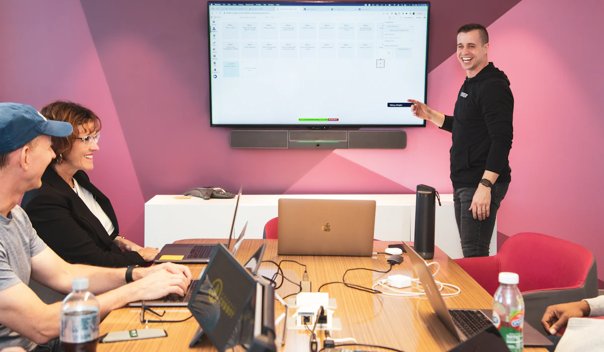 A trainer presenting to a laptop-equipped team gathered around a table, pointing at a wall-mounted screen