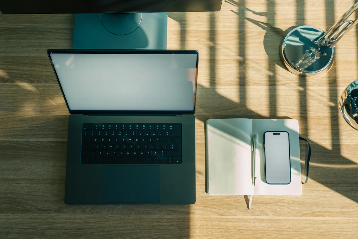 A laptop, open notebook and phone on a wooden desk in morning light