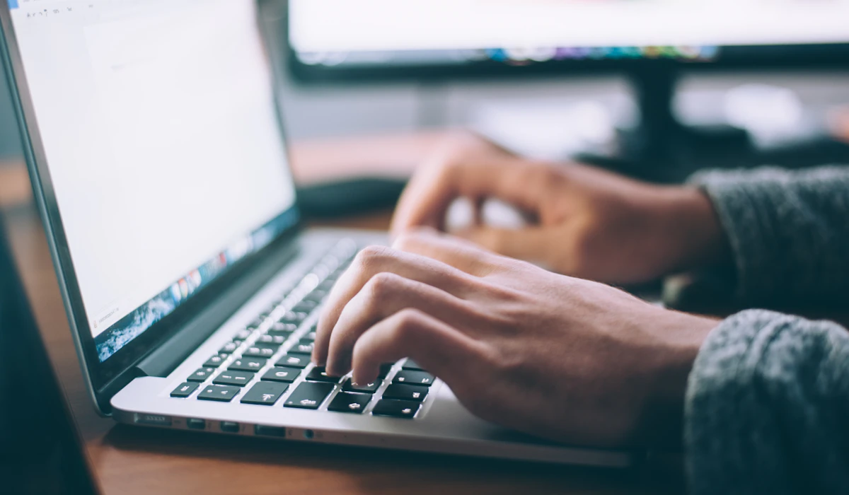 Close-up of hands typing on a laptop keyboard at a desk with a second monitor in the background