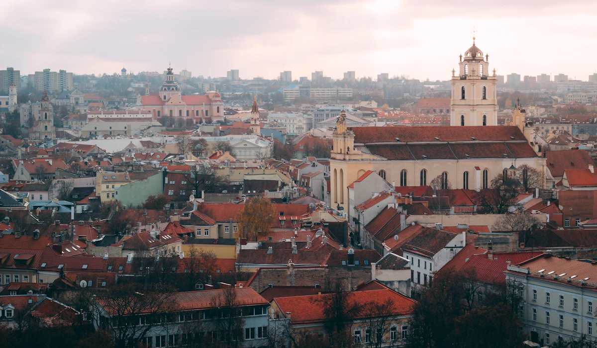 A high view over Vilnius Old Town at dusk, red-tiled roofs running to the horizon with a tall white baroque bell tower standing over them