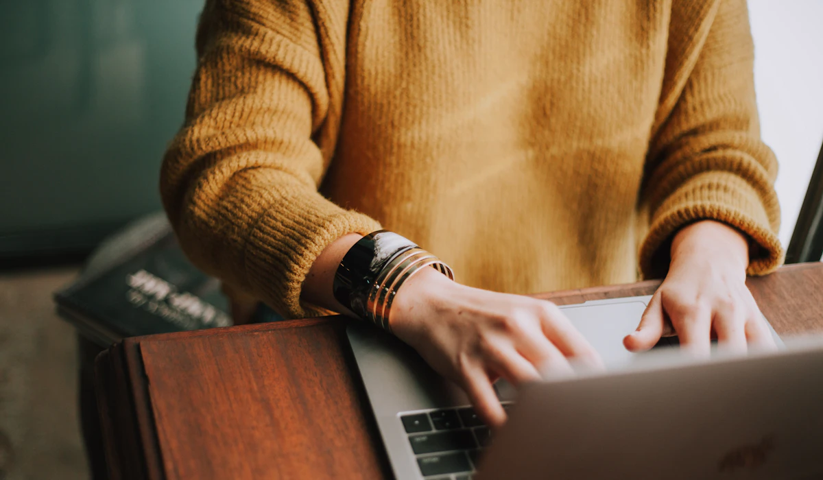 A person in a mustard sweater typing on a laptop at a wooden desk